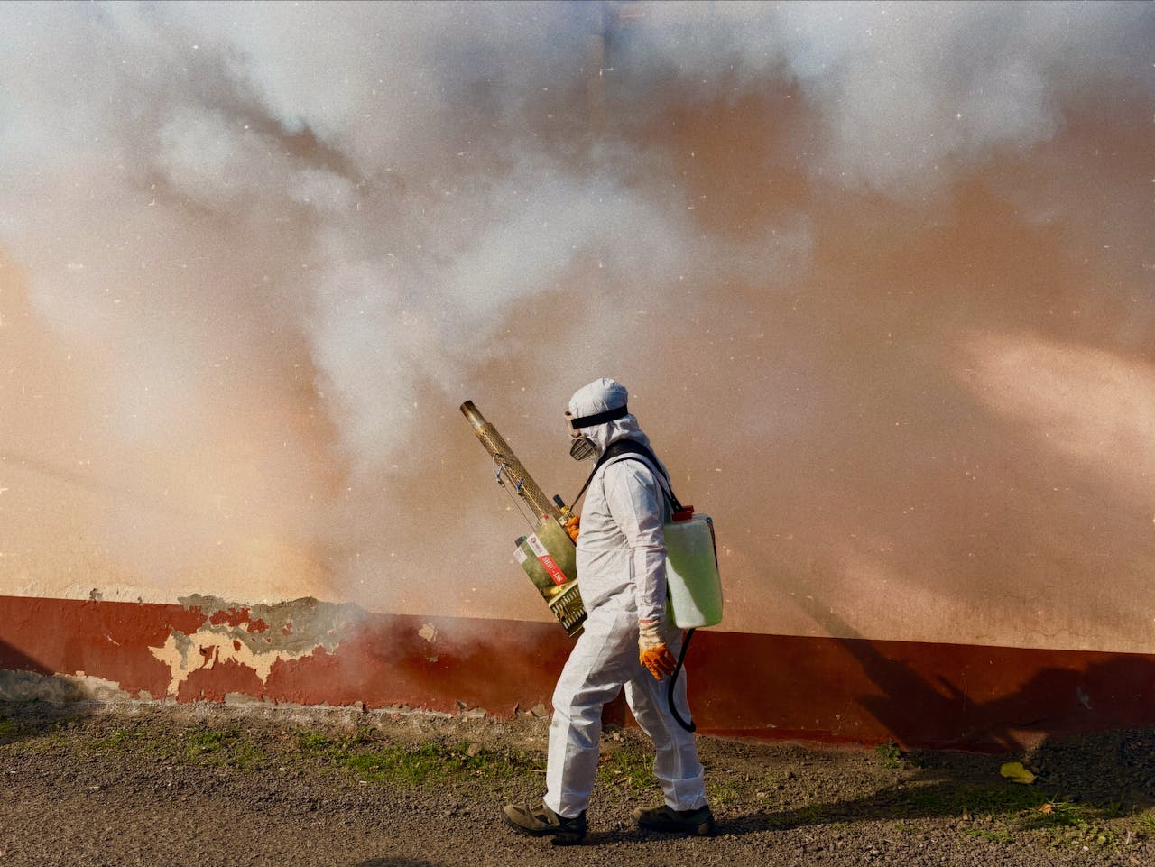 services-03 A worker in a full protective suit fumigates an area outdoors, spraying pesticide.